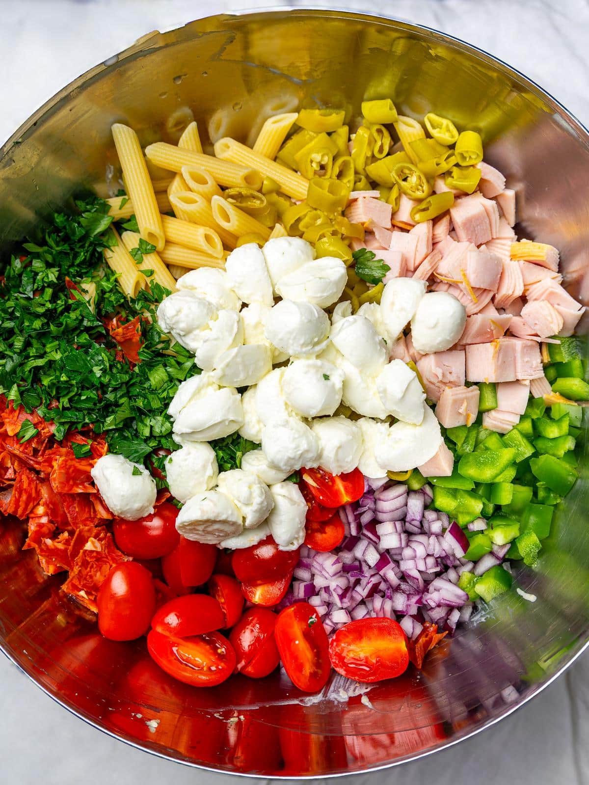 Unmixed italian grinder pasta salad ingredients with penne, mozzarella, turkey, tomatoes, peppers, and herbs