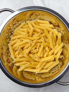 Cooked penne pasta draining in a metal colander on a marble counter