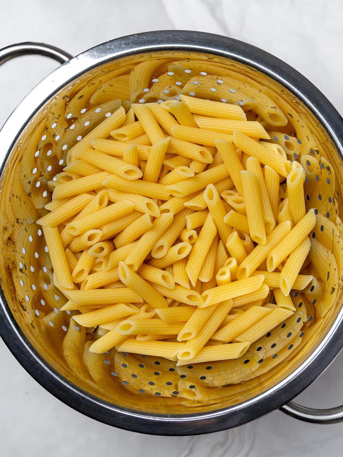 Cooked penne pasta draining in a metal colander on a marble counter