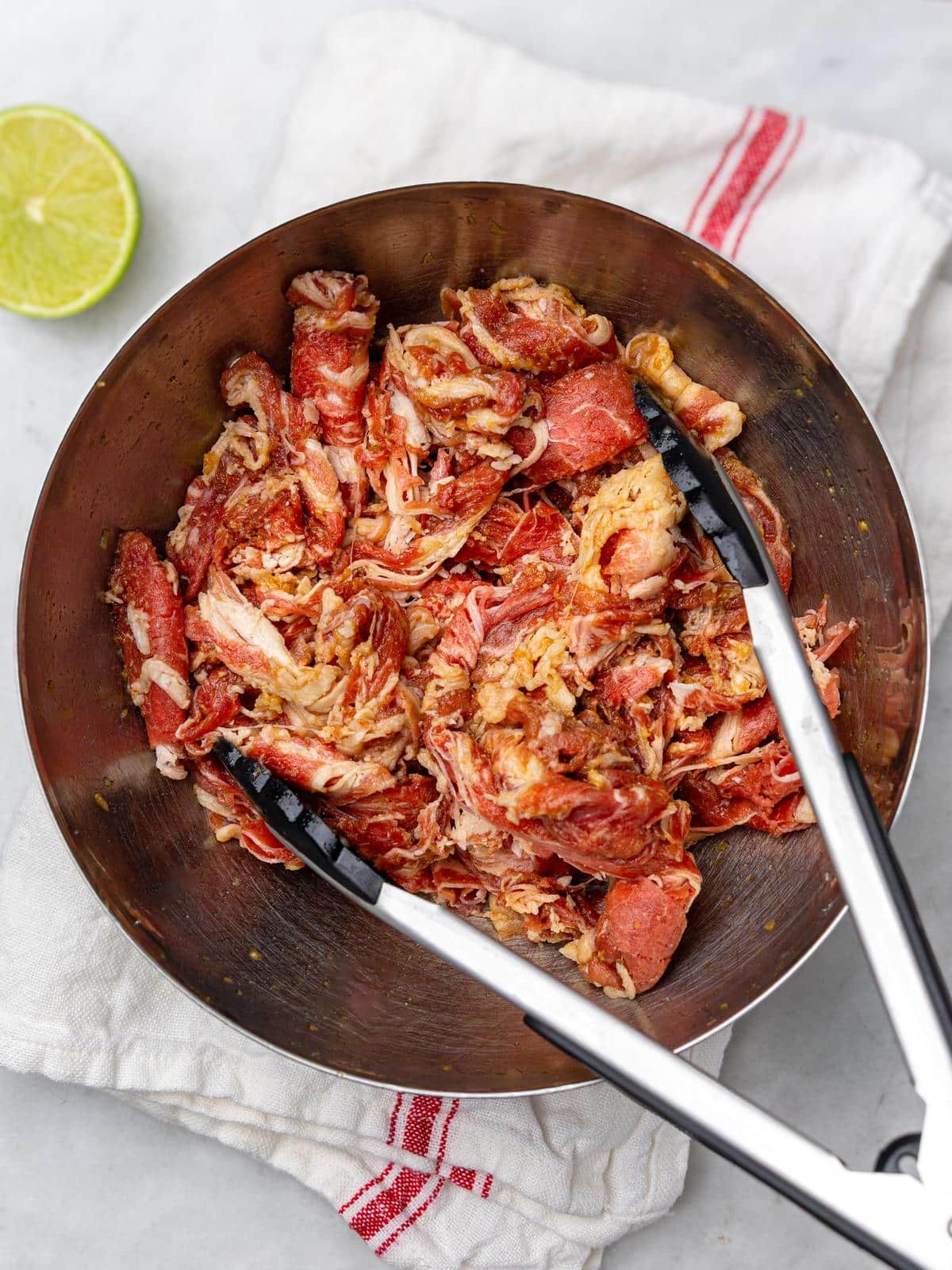 Thinly sliced marinated beef in a metal bowl with tongs and a lime half