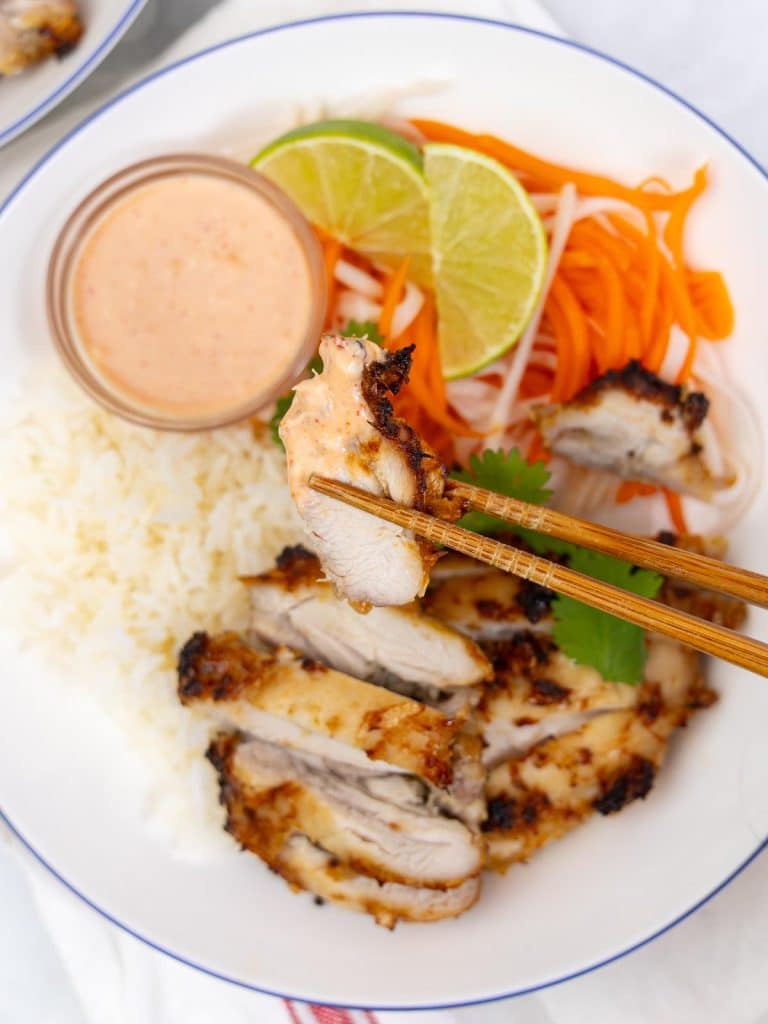 Chopsticks lifting a piece of charred lemongrass chicken dipped in sriracha mayo over a rice bowl