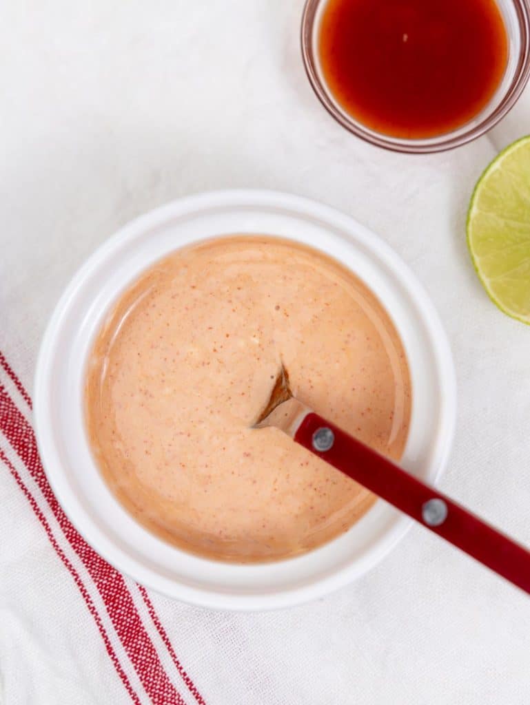 Creamy sriracha mayo in a small white ramekin with sriracha and a lime wedge on the side