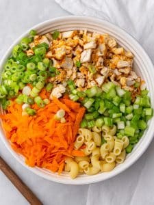 Chopped chicken, celery, shredded carrots, green onions and pasta in a white bowl