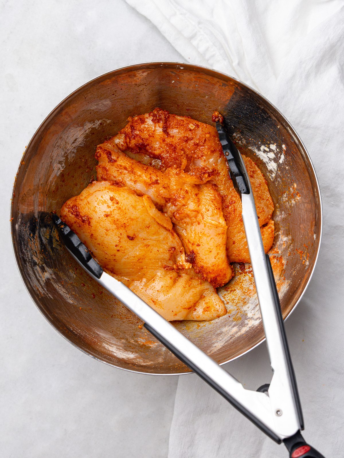 Chicken breasts coated in seasoning in a metal mixing bowl with tongs