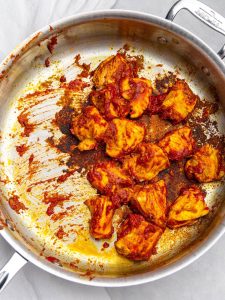 Chicken pieces searing with red curry paste and tomato paste in a stainless steel skillet
