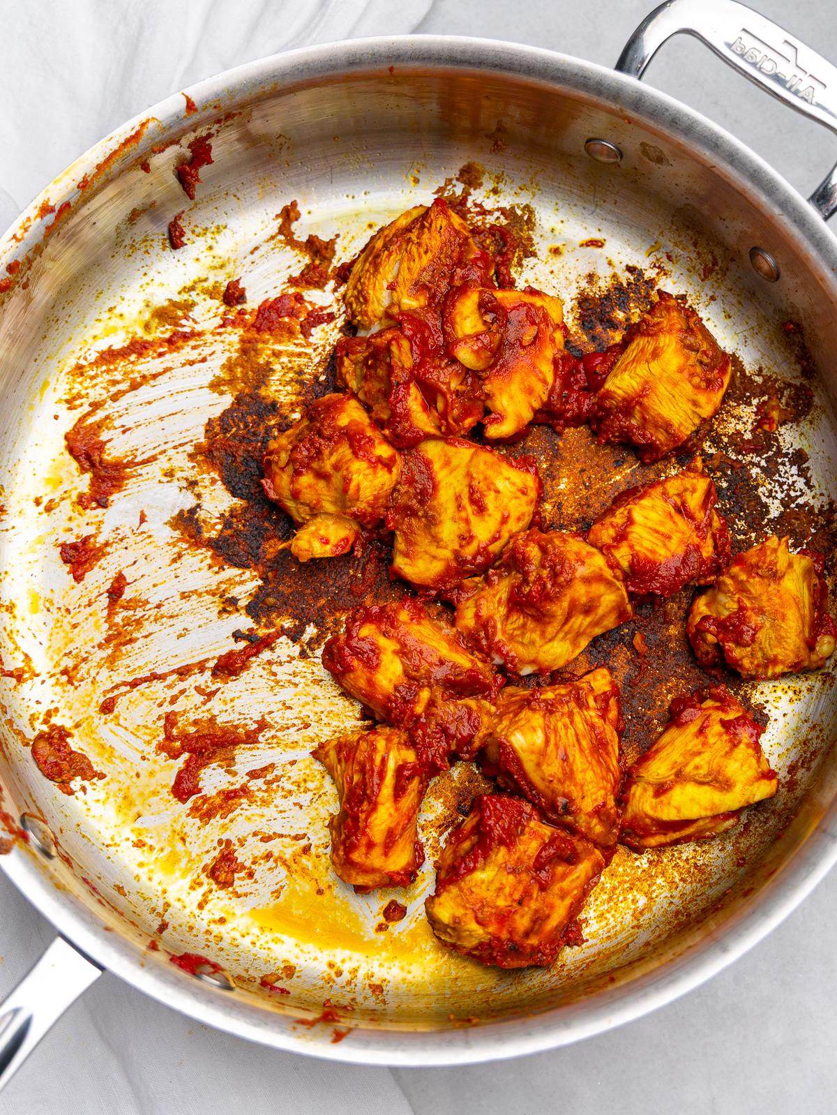 Chicken pieces searing with red curry paste and tomato paste in a stainless steel skillet