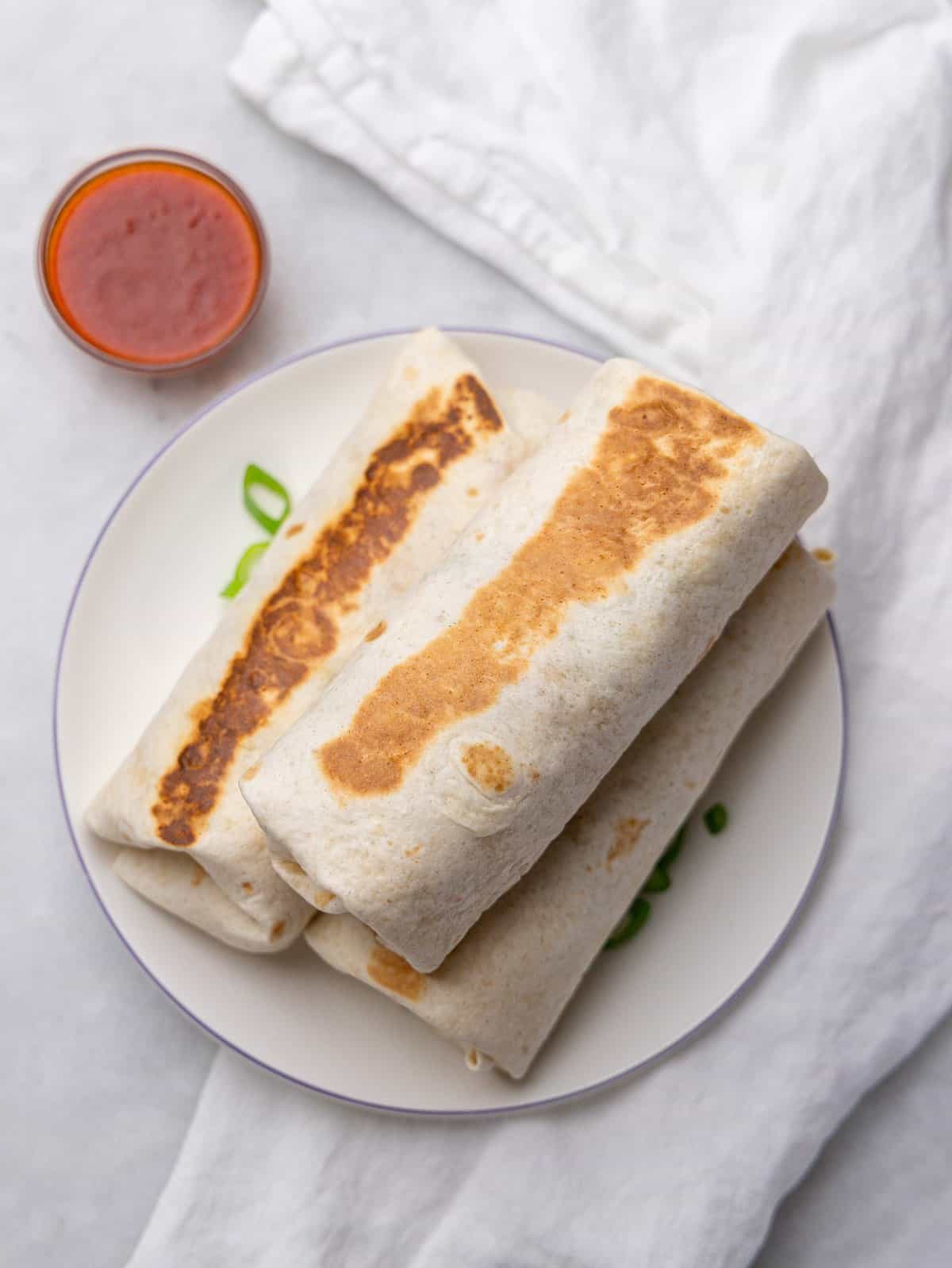Golden toasted healthy breakfast burritos stacked on a plate with hot sauce