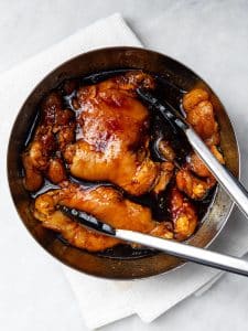 Chicken thighs marinating in orange teriyaki marinade in a metal bowl with tongs