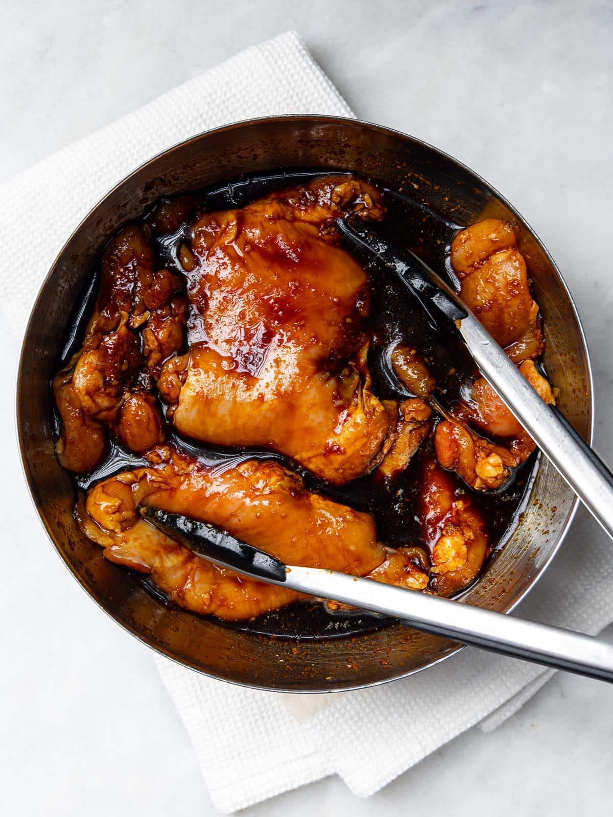 Chicken thighs marinating in orange teriyaki marinade in a metal bowl with tongs