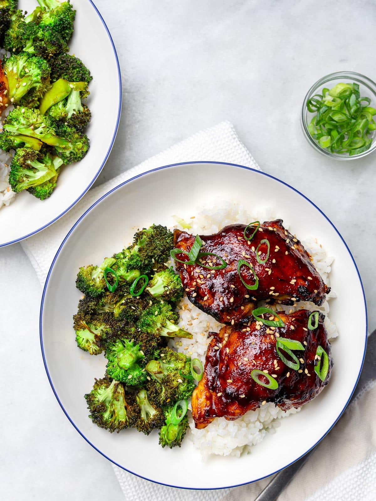 Two teriyaki chicken rice bowls with crispy broccoli and a small bowl of sliced green onions