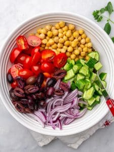 Chickpeas, cherry tomatoes, cucumber, kalamata olives, and red onion arranged in a white bowl before tossing
