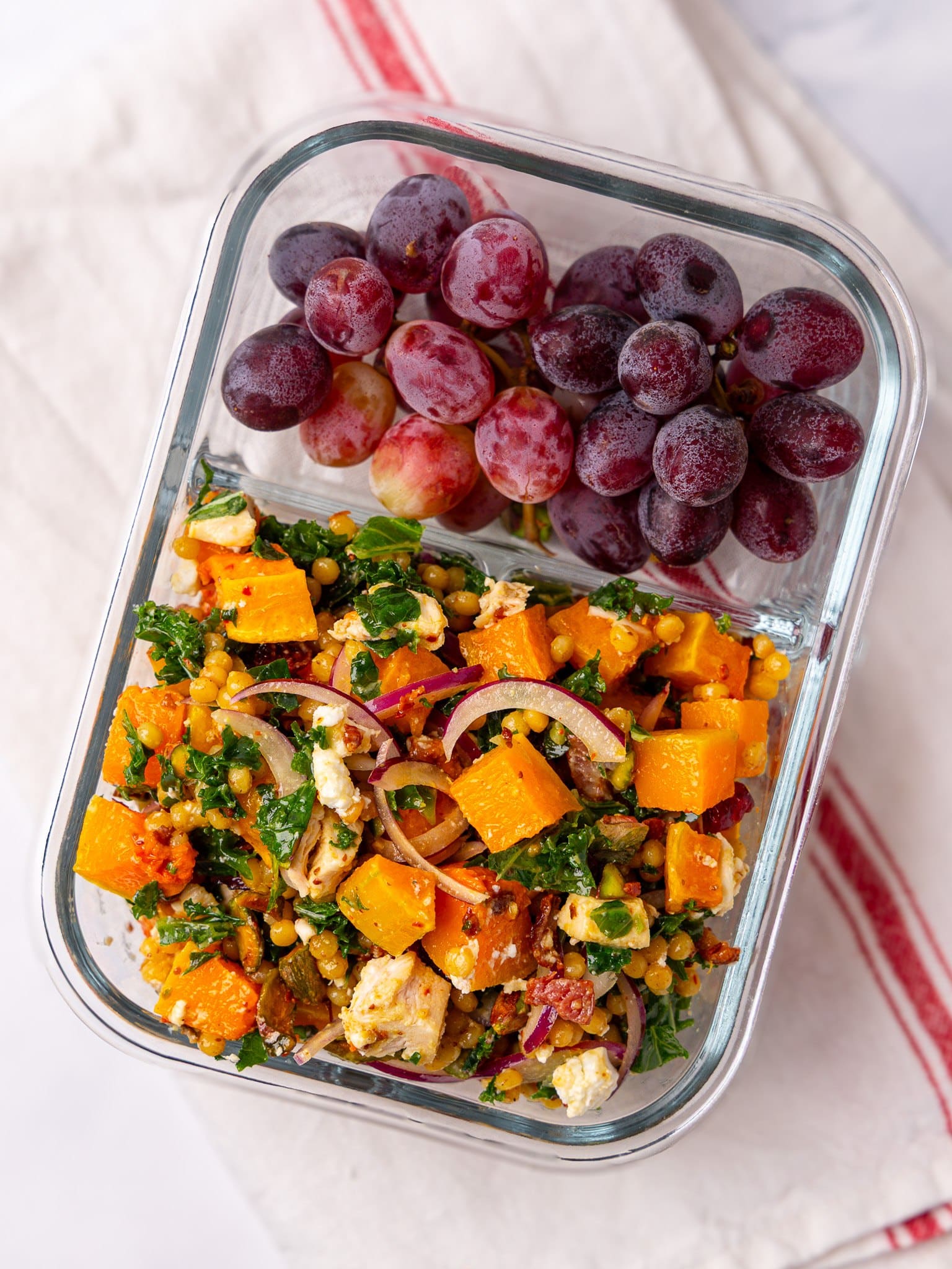 Autumn couscous salad meal prep in a glass container with red grapes on the side
