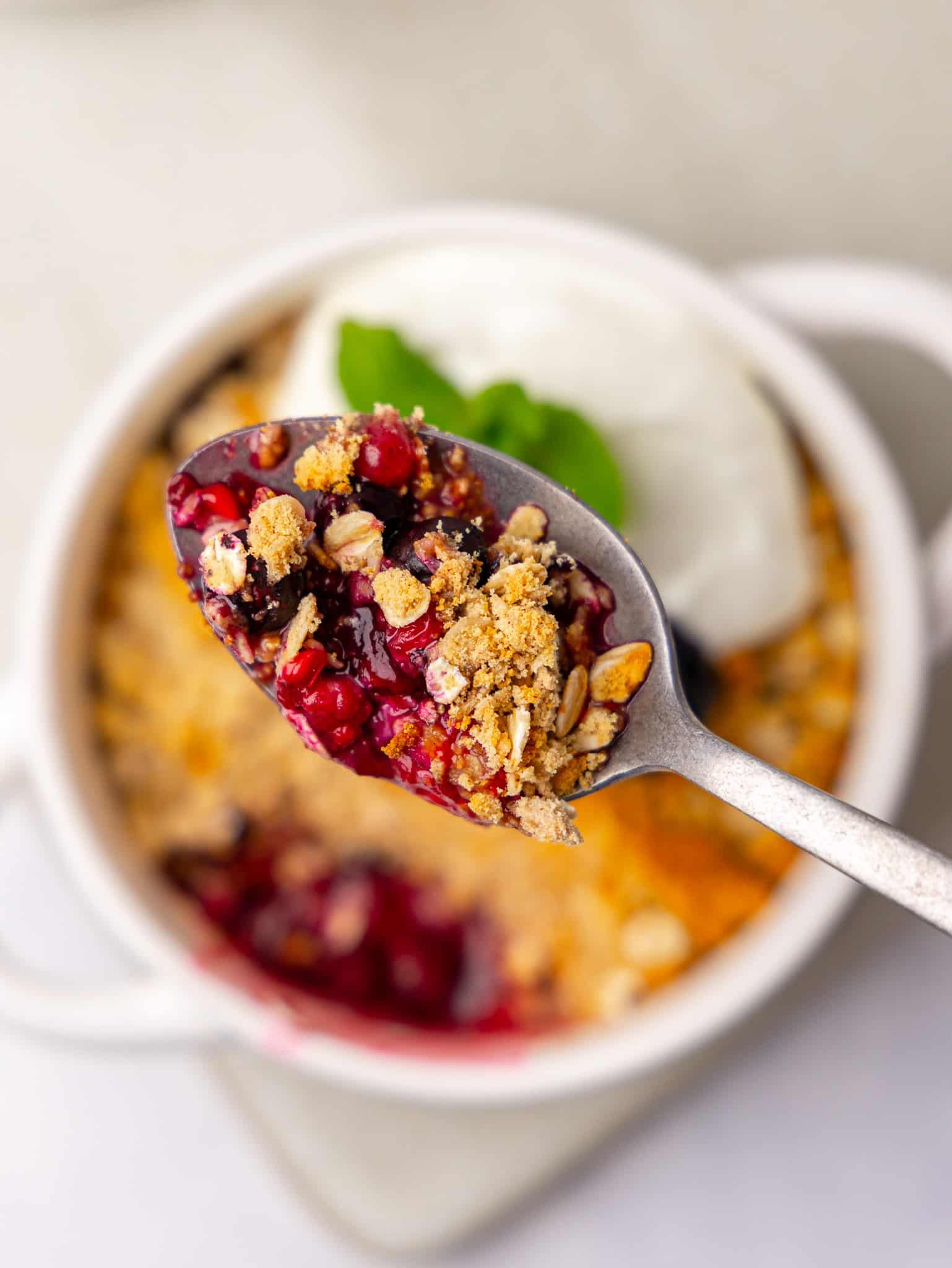 Close-up of a spoon of berries and a healthy crumble topping with oats over a ramekin of greek yogurt