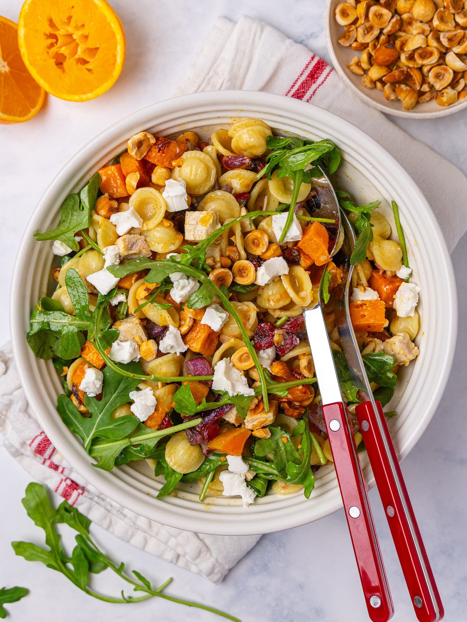 Fall pasta salad with butternut squash, arugula, goat cheese and hazelnuts in a white bowl with red serving spoons