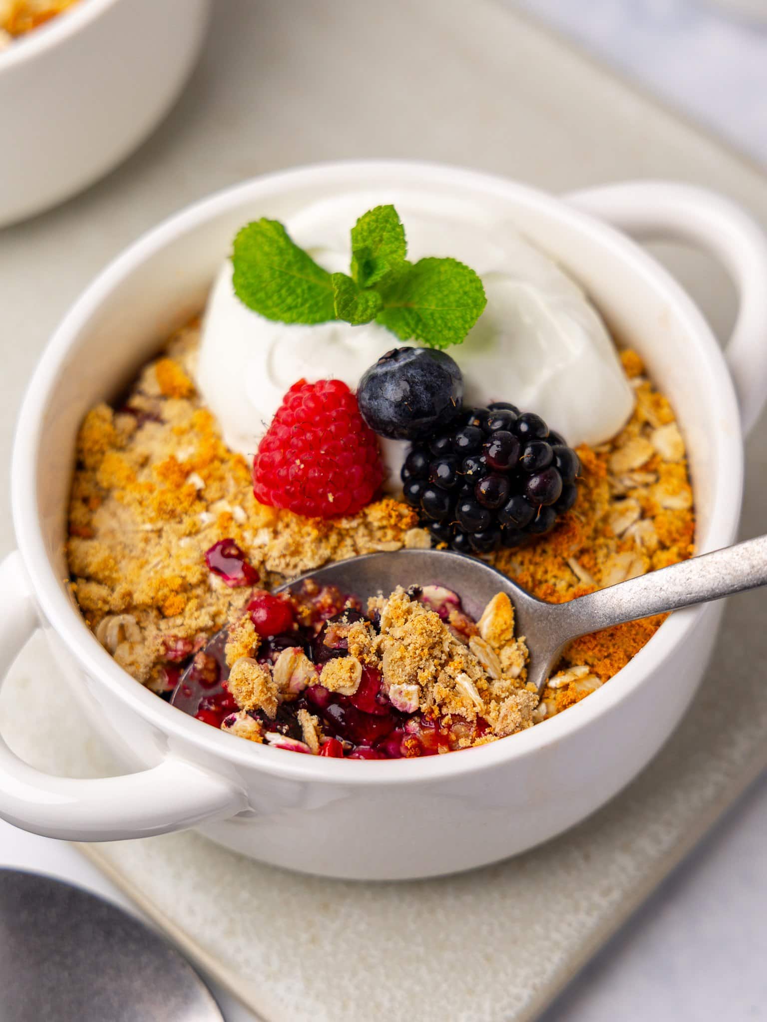 High protein berry crumble in a white ramekin with a spoon scooping into the jammy mixed berry filling under greek yogurt and mint