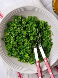Massaged chopped kale in a white bowl with salad servers