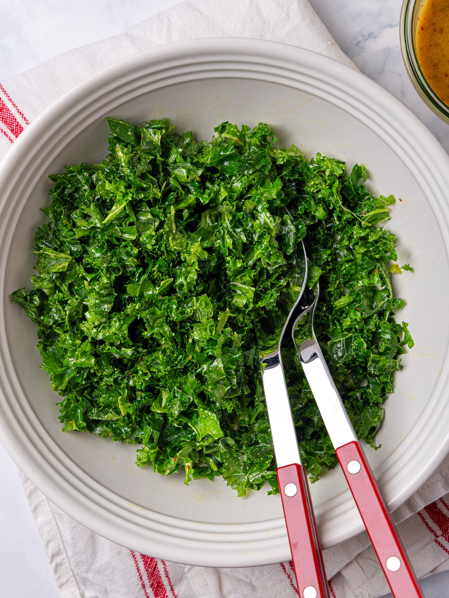 Massaged chopped kale in a white bowl with salad servers