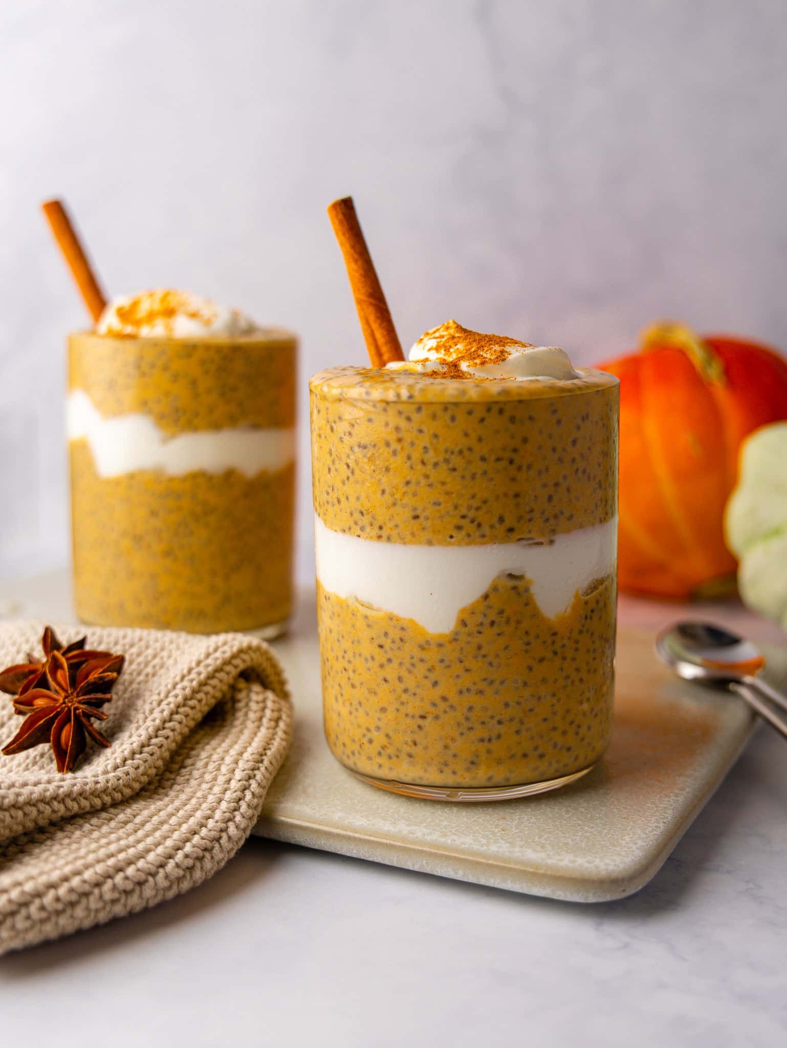 Two glasses of pumpkin spice chia pudding with cinnamon sticks, star anise and a pumpkin behind